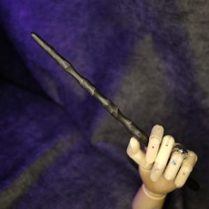 Puede incluir: Una varita marrón oscuro con un diseño texturizado y acanalado. La varita está sujeta por una mano de madera, con un anillo en un dedo. El fondo es una mezcla de morado y gris.