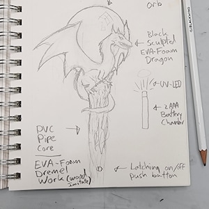 May include: A hand-drawn sketch of a dragon sculpture with a translucent frosted white orb on top. The dragon is made of black sculpted EVA foam and is mounted on a PVC pipe core. The sculpture is lit by a UV LED and powered by two AAA batteries. The sketch includes notes about the materials and construction of the sculpture.