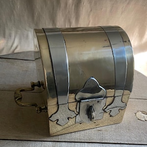 May include: A small, antique-style metal chest with a curved top. The chest is made of brass and silver-colored metal, with decorative accents and a latch closure. It features a handle on the side and is likely used for storing small items.