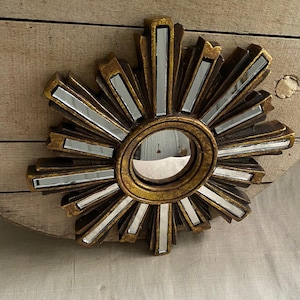 May include: A gold-colored sunburst mirror with a small circular mirror at the center. The sunburst design features alternating gold and mirrored segments radiating outwards. The mirror is a decorative wall hanging.