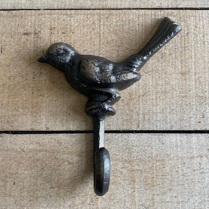 Op de afbeelding: Een gietijzeren haak in de vorm van een vogel met een donker bronzen afwerking. De vogel zit boven een haak, ontworpen om items op te hangen. De haak is gemonteerd op een houten oppervlak.