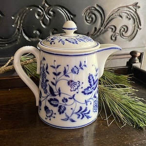 Puede incluir: Una tetera de cerámica blanca y azul con tapa. La tetera presenta un patrón floral azul y un asa curva. El pico está en el lado derecho. La tetera está sobre una superficie de madera oscura.