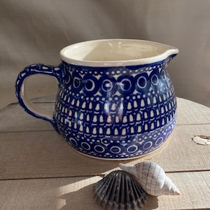 May include: A blue and white ceramic pitcher with a decorative pattern. The pitcher has a rounded body and a handle. It is sitting on a wooden surface with a seashell.