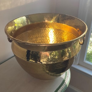 Puede incluir: Un macetero grande de latón martillado con dos asas decorativas. El macetero está sobre una mesa blanca y verde.