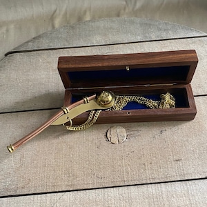 Puede incluir: Un silbato de latón con mango de cobre y cadena dorada, en una caja de madera con forro de terciopelo azul.