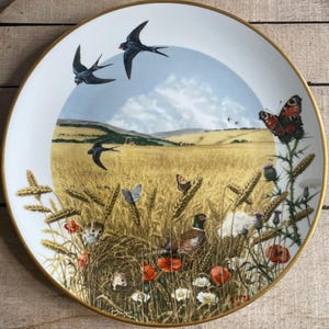 May include: A decorative plate with a gold rim features a pastoral scene. The image includes swallows in flight, butterflies, wheat, poppies, and a pheasant. The background depicts a field and a blue sky.