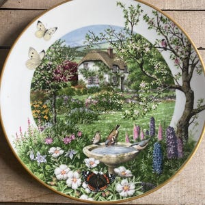 May include: A decorative plate featuring a detailed garden scene with a cottage, birds, butterflies, and flowers. The plate has a gold rim and a white background. The image depicts a serene outdoor setting.