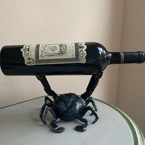 May include: A decorative wine bottle holder in the shape of a crab. The crab is made of metal with a blue-green finish. A dark wine bottle rests horizontally in the crab's claws. The bottle has a label with text.