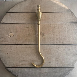 May include: A brass fireplace tool with a decorative handle and a curved hook. The tool is set against a round, light-colored wooden background. The hook is designed for handling hot items.