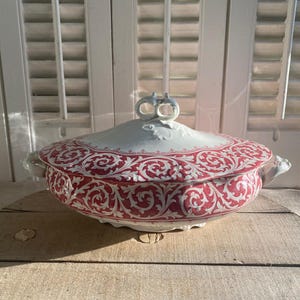 Puede incluir: Un plato de cerámica blanco y rojo con tapa. El plato presenta un detallado patrón floral rojo alrededor del borde y en la tapa. Las asas son pequeñas y curvas. El plato está sobre una superficie de madera.