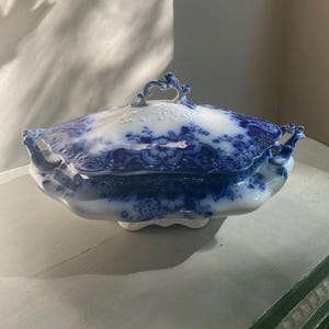 Puede incluir: Un plato de porcelana azul y blanco con tapa. El plato tiene un estampado floral y asas ornamentadas. La tapa presenta un asa decorativa. El plato está sobre una superficie clara.