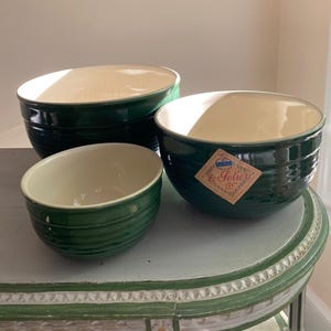 May include: Three dark green ceramic mixing bowls with cream-colored interiors and horizontal ribbed detailing on the exterior. One bowl features a diamond-shaped paper label with the text le Sotier printed in a decorative script font.