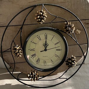 May include: A round wall clock with a black metal frame featuring five decorative gold-toned pinecones attached to the outer ring. The clock face is cream-colored with black Roman numerals and the word QUARTZ printed in the center.