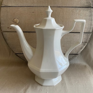 May include: A white ceramic teapot with a lid and a handle. The teapot has a rounded body and a square base.