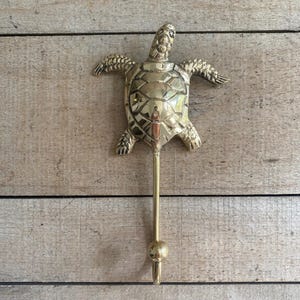 May include: A brass turtle wall hook. The hook is shaped like a turtle with outstretched flippers and a textured shell. The hook extends downward with a rounded end. The hook is mounted on a wooden background.
