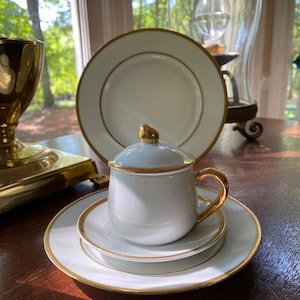 Puede incluir: Un juego de té de porcelana blanca con ribete dorado. El juego incluye una taza con tapa, dos platillos y un plato llano. La taza tiene un asa dorada y un adorno dorado en la tapa. Los platos tienen un borde dorado.