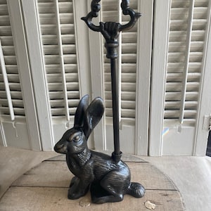 Puede incluir: Un tope de puerta de metal negro con una detallada escultura de conejo. El tope de puerta tiene una varilla larga y delgada con un asa ornamentada. El conejo está sentado sobre una superficie de madera, con un fondo de contraventanas blancas.