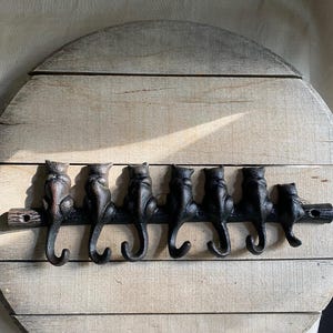 Può includere: Un attaccapanni rotondo in legno con sei ganci neri a forma di gatto. I gatti sono appollaiati su un ramo orizzontale, aggiungendo un tocco stravagante al design funzionale. Lo sfondo in legno ha una finitura chiara e naturale.