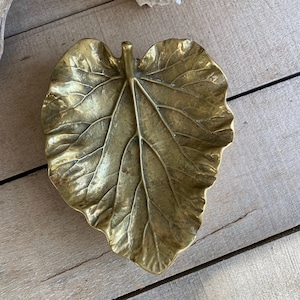 Puede incluir: Un plato en forma de hoja de latón con vetas detalladas. El plato tiene un tono dorado y descansa sobre una superficie de madera. Una concha blanca está en el fondo. Esta pieza decorativa es ideal para guardar objetos pequeños.