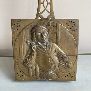 May include: Antique brass fireplace tool with a decorative handle and a square panel featuring a raised image of a man smoking a pipe. The panel has a decorative border and is perforated with small holes.