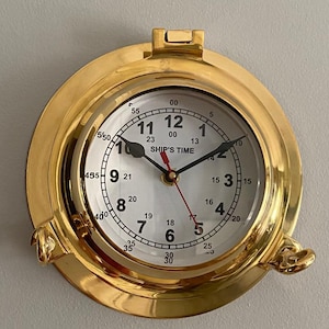 May include: A brass-colored, round wall clock with a white face and black numerals. The clock has a nautical porthole design with the words "SHIP'S TIME" printed on the face. The clock is mounted on a gray wall.