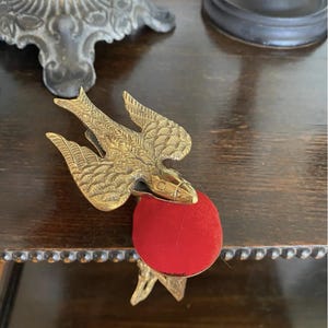May include: A brass bird-shaped sewing clamp featuring detailed wing engravings and a circular red velvet pincushion attached to the front. The metal bird is positioned with its wings spread, serving as a functional tool for holding fabric.