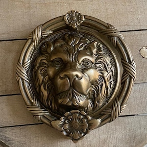 May include: A gold lion head door knocker with a decorative floral design. The knocker is mounted on a wooden surface.