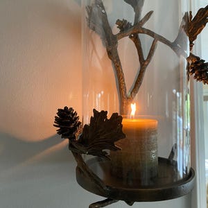 Op de afbeelding: Een decoratieve kandelaar met een brandende kaars in een heldere glazen cilinder. De houder is versierd met bronskleurige dennenappels, bladeren en takken. De kaars is olijfgroen en getextureerd.