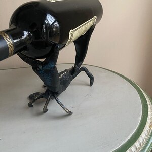 May include: A black wine bottle rests in a crab-shaped metal wine holder. The crab is dark with blue-green accents. The bottle has a label and a gold design near the top. The holder sits on a round table.