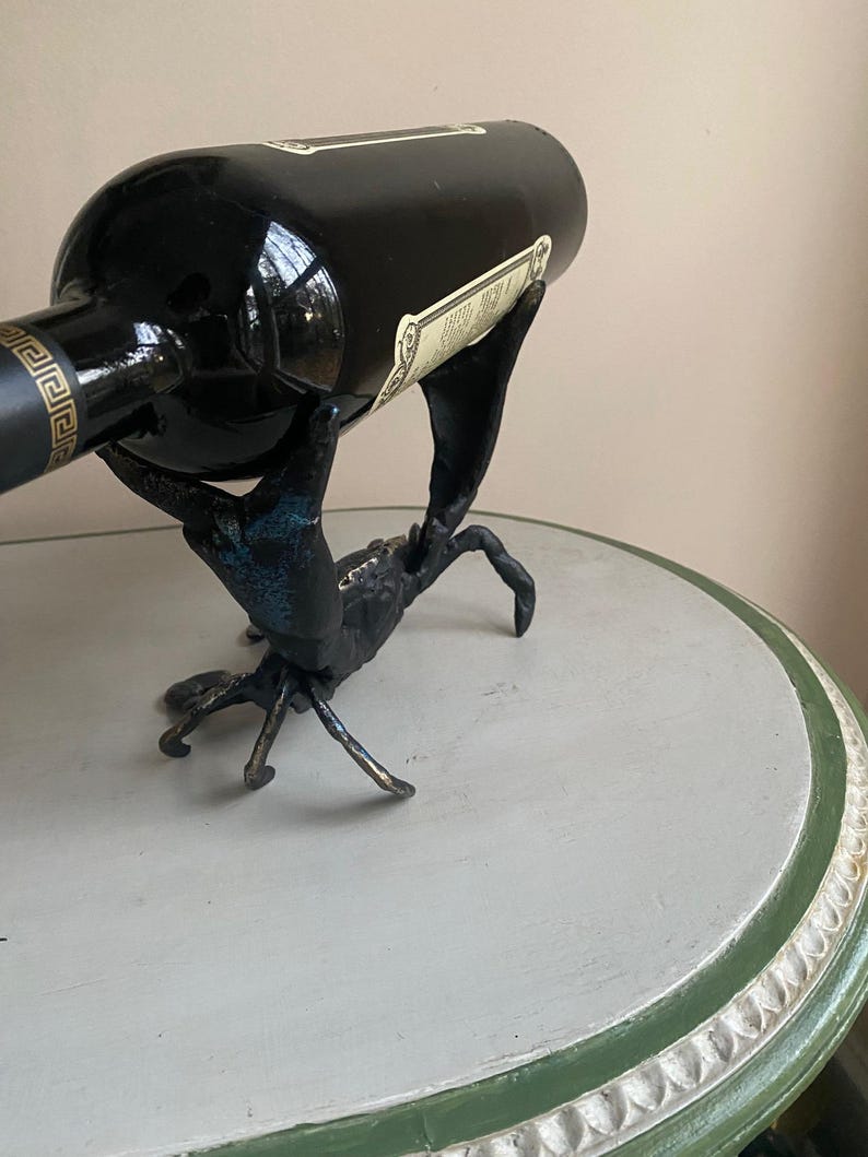 May include: A black wine bottle rests in a crab-shaped metal wine holder. The crab is dark with blue-green accents. The bottle has a label and a gold design near the top. The holder sits on a round table.