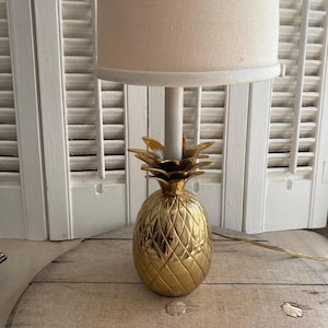 Puede incluir: Una lámpara de mesa dorada con forma de piña y una pantalla de color crema. La lámpara tiene una superficie texturizada y un tallo blanco. La lámpara está sobre una superficie de madera frente a contraventanas blancas.