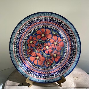 Op de afbeelding: Een grote, ronde, blauw-witte keramische plaat met een bloemenmotief. De plaat heeft een rand van blauw-witte geometrische patronen, en het midden van de plaat is versierd met een cluster van rode bloemen met groene bladeren.
