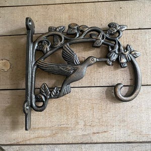 May include: A cast iron hummingbird bracket in a bronze finish. The bracket features a hummingbird in flight, feeding from a flower, with leaves and vines. The bracket has a hook for hanging items and a flat plate for mounting.
