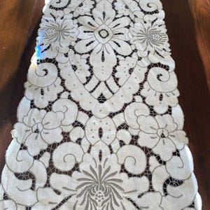 May include: White linen table runner with a delicate, intricate floral design embroidered in silver thread. The design features a central motif of a large flower with smaller flowers and leaves surrounding it.