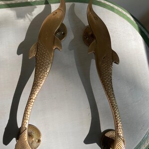 May include: A pair of gold-toned metal door handles shaped like dolphins. Each handle features a textured scale pattern along the body, a smooth head, and a flared tail fin. The handles are mounted on circular bases with visible screw holes.