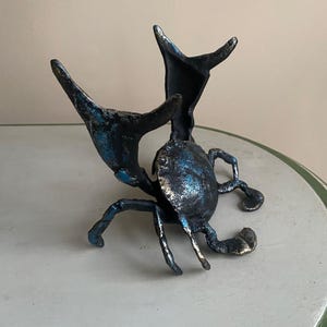May include: A bronze crab sculpture with blue and silver accents. The crab has outstretched claws and legs, and is positioned on a light-colored surface. The sculpture is a decorative item.