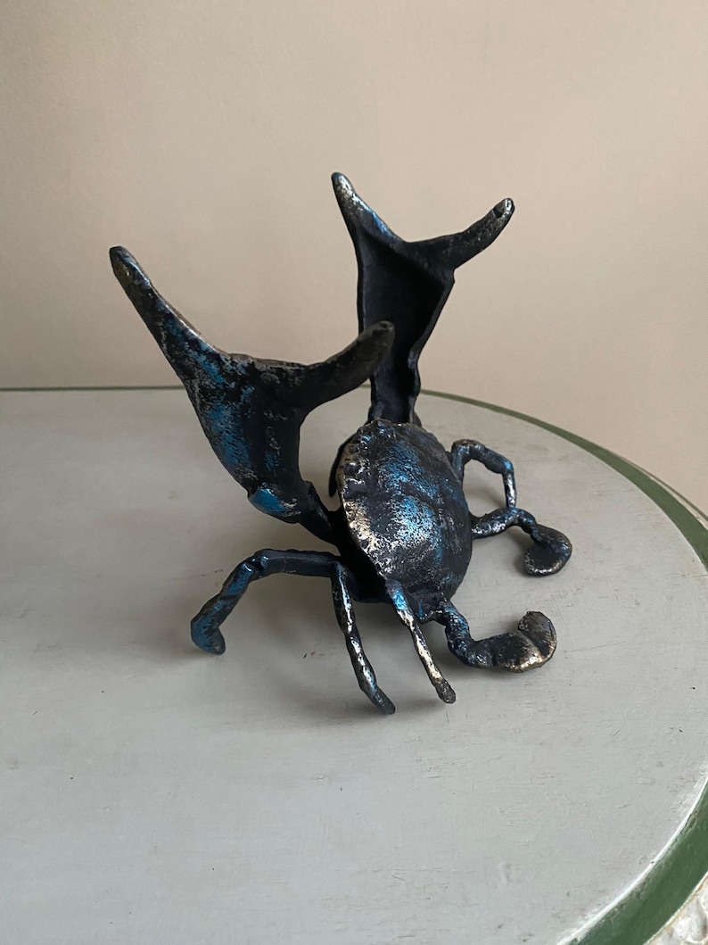 May include: A bronze crab sculpture with blue and silver accents. The crab has outstretched claws and legs, and is positioned on a light-colored surface. The sculpture is a decorative item.