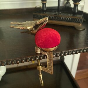 May include: A brass sewing clamp featuring a bird-shaped top and a bright red velvet pincushion. The metal clamp has an engraved floral pattern and a screw mechanism for attaching to a table edge, providing a functional tool for sewing tasks.