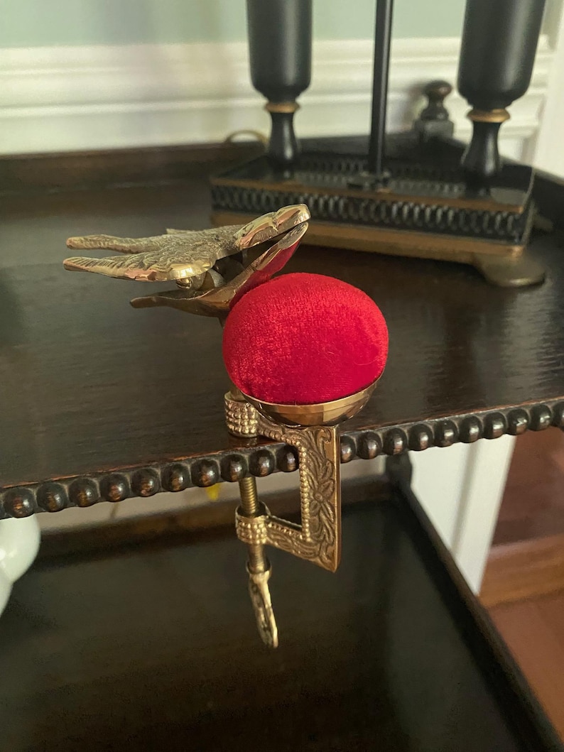 May include: A brass sewing clamp featuring a bird-shaped top and a bright red velvet pincushion. The metal clamp has an engraved floral pattern and a screw mechanism for attaching to a table edge, providing a functional tool for sewing tasks.