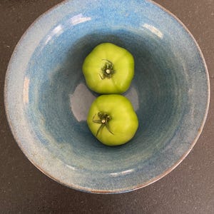 Könnte beinhalten: Zwei grüne Tomaten in einer blauen Keramikschüssel. Die Tomaten sind unreif und haben einen grünen Stiel.