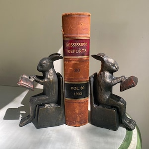 May include: A pair of dark bronze-toned metal bookends, each shaped like a rabbit sitting on a block and holding a small book. Between the two rabbit bookends stands an antique, orange-brown leather-bound book titled Mississippi Reports Vol. 80 1902.