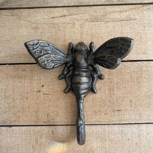 May include: A cast iron bee-shaped wall hook with textured wings and body. The hook is dark gray with bronze highlights, and is mounted on a light brown wooden surface. The bee's wings are spread, and the hook extends downward.
