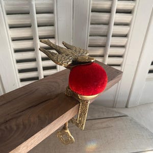May include: A brass pin cushion clamp featuring a bird figure with outstretched wings mounted atop a vibrant red velvet cushion. The metal base includes an ornate screw mechanism with a key-shaped handle designed to attach to a flat surface edge.