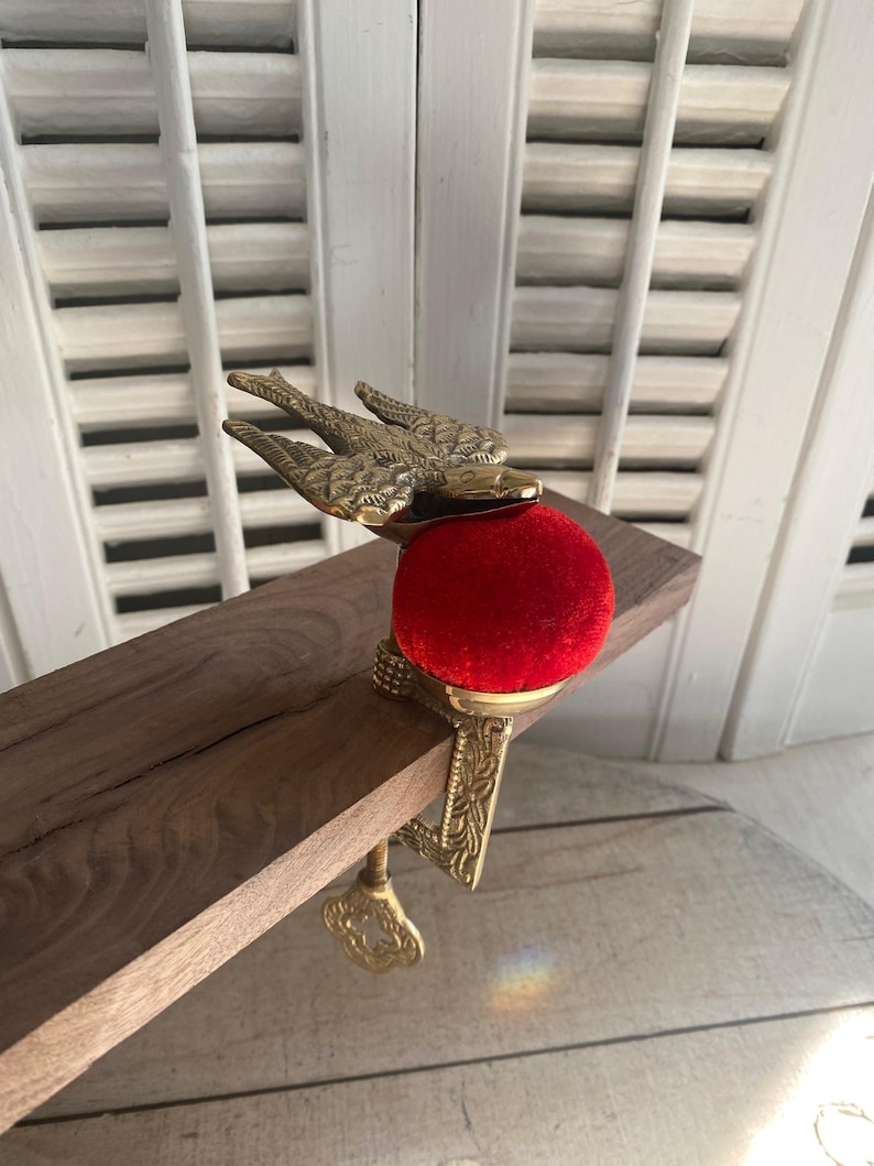 May include: A brass pin cushion clamp featuring a bird figure with outstretched wings mounted atop a vibrant red velvet cushion. The metal base includes an ornate screw mechanism with a key-shaped handle designed to attach to a flat surface edge.