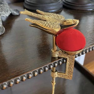 May include: A brass sewing clamp featuring a bird figure with outstretched wings attached to a circular red velvet pincushion. The metal clamp has an engraved floral pattern and a screw mechanism for securing it to a flat surface edge.