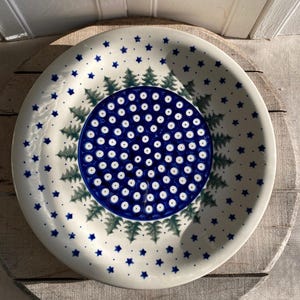 May include: White ceramic plate with a blue and green Christmas tree design. The plate features a central blue circle with white dots, surrounded by green tree motifs and blue star accents around the rim.