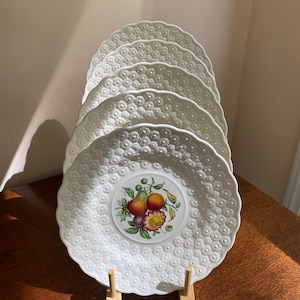 May include: Four white ceramic plates with a raised embossed design and a floral pattern with fruit in the center. The plates are stacked on a wooden stand.