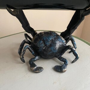 May include: A crab-shaped wine bottle holder. The sculpture is made of metal with a blue and bronze finish. The crab's claws extend upwards to cradle a wine bottle. The crab's body is textured, and the legs are bent in a walking position.