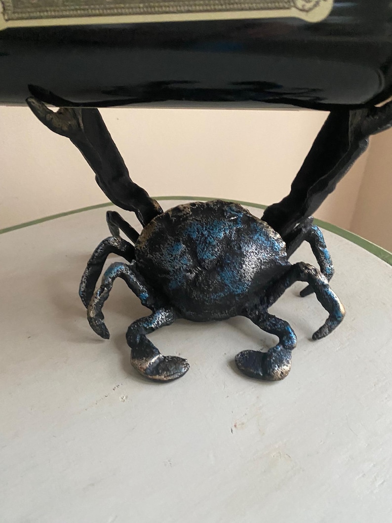 May include: A crab-shaped wine bottle holder. The sculpture is made of metal with a blue and bronze finish. The crab's claws extend upwards to cradle a wine bottle. The crab's body is textured, and the legs are bent in a walking position.