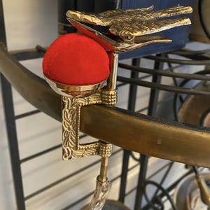 May include: A brass sewing clamp featuring an embossed floral pattern on the vertical arm and a detailed bird figure at the top. A bright red, spherical fabric pincushion is mounted in a polished brass cup attached to the side of the metal clamp.